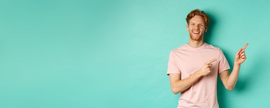 Handsome male model with red messy hair showing advertisement on copy space, pointing at upper right corner and smiling happy, standing over turquoise background.