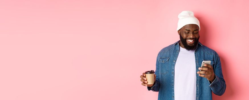 Image of stylish Black man hipster drinking takeaway coffee, reading message on phone and smiling, standing over pink background.