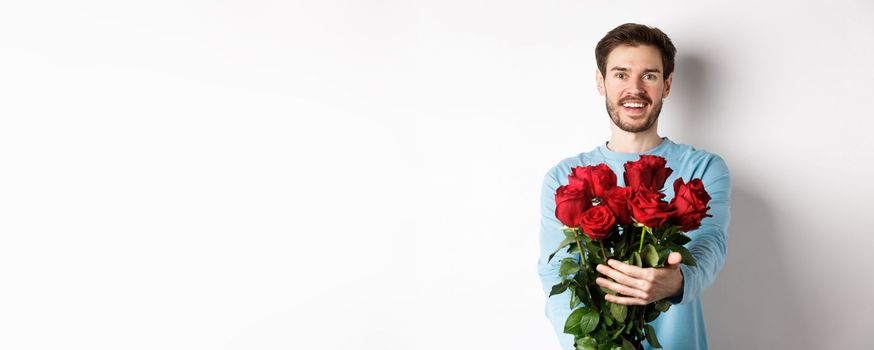 Handsome bearded guy stretch out hands, giving bouquet of roses and smiling, bring flowers on romantic date, celebrating Valentines day with lover, standing over white background.