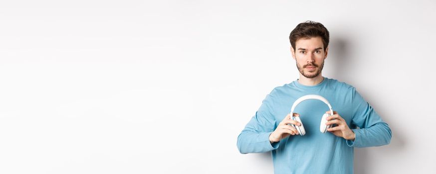 Young bearded guy in blue sweatshirt put on wireless headphones, listening music, standing on white background.