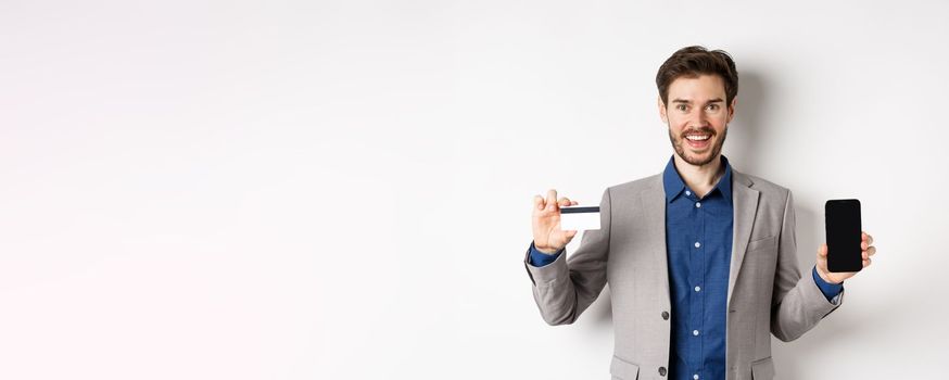 Online shopping. Smiling business man in suit showing plastic credit card with empty smartphone screen, standing against white background.