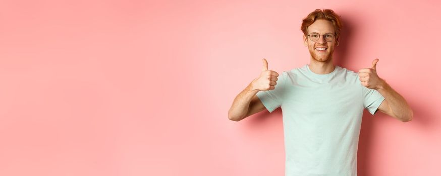 Cheerful european man with red hair and beard, wearing glasses, showing thumbs-up and smiling in approval, praise something good, standing over pink background.