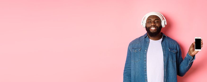 Happy hipster guy listening music on headphones and showing mobile screen, smiling satisfied, standing over pink background.