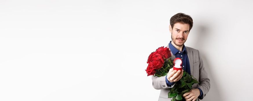 Romantic man with boquet of red roses asking to marry him, holding engagement ring and looking confident at camera, standing in suit over white background.