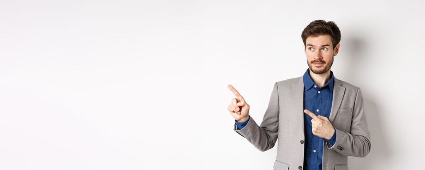 Doubtful male entrepreneur in suit looking and pointing left with hesitant face, standing unsure against white background.