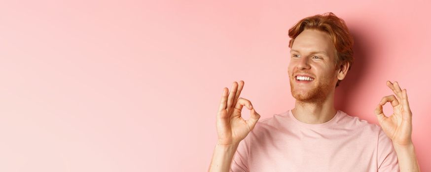 Close up of handsome redhead man in t-shirt, smiling satisfied and looking left, showing okay signs in approval, praising awesome product, standing over pink background.