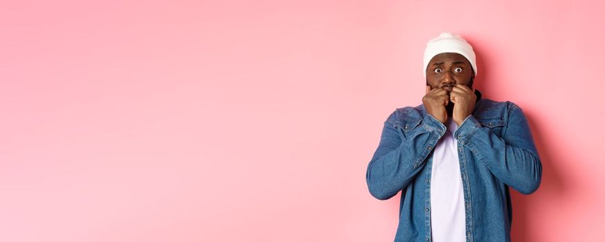 Scared Black guy trembling from fear, biting fingernails and staring at camera, standing over pink background.