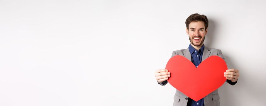 Handsome young man wishing happy Valentines day, giving big red heart sign and smiling, make surprise to lover, standing in suit over white background.