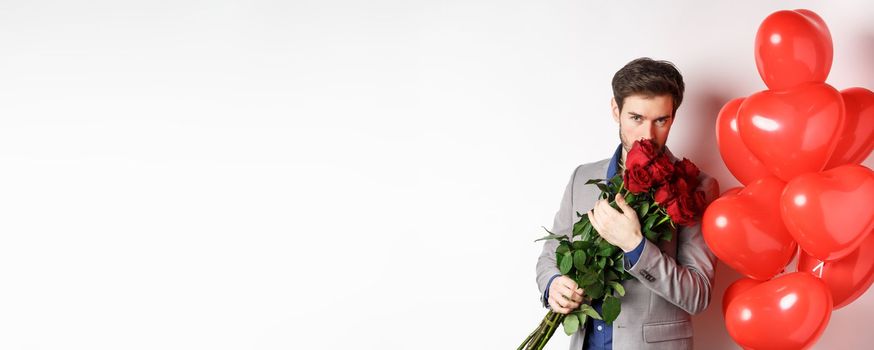 Romantic man smell bouquet of red roses and looking passionate at camera. Boyfriend in suit going on Valentines date with gifts and heart balloons, white background.