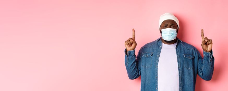 Coronavirus, lifestyle and global pandemic concept. Upset Black man in face mask frowning, pointing fingers up, showing advertisement, standing over pink background.