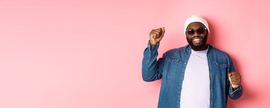 Happy Black man in beanie an sunglasses rejoicing, dancing with happy face, standing over pink background.