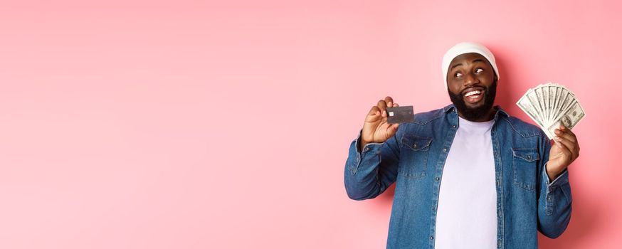 Dreamy african-american man showing credit card and dollars, thinking about shopping and smiling, standing over pink background.