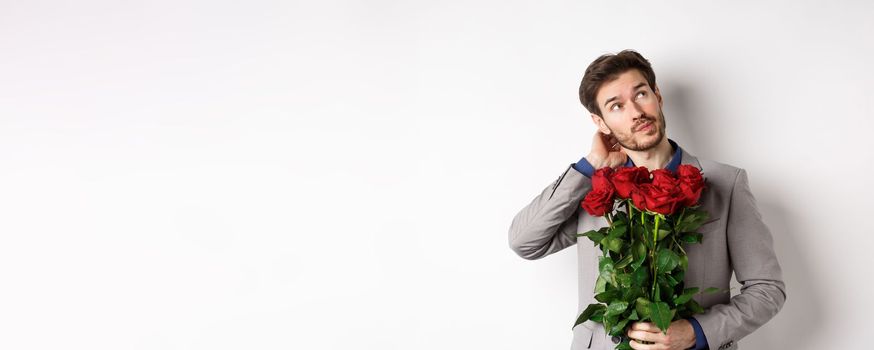 Thoughtful young man in suit going on valentines day date, holding bouquet of roses, thinking and scratching head, standing against white background.