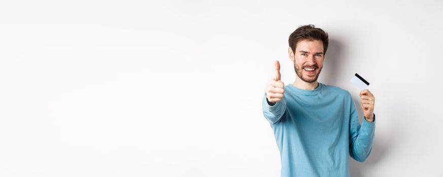 Cheerful young man showing plastic credit card and thumb up, like and approve good bank service, standing over white background.