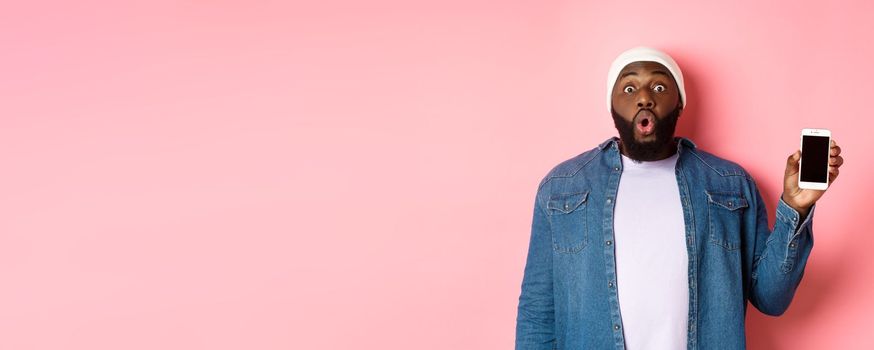 Amazed Black guy in streetstyle clothes showing mobile phone screen, saying wow as demonstrating smartphone display, standing over pink background.