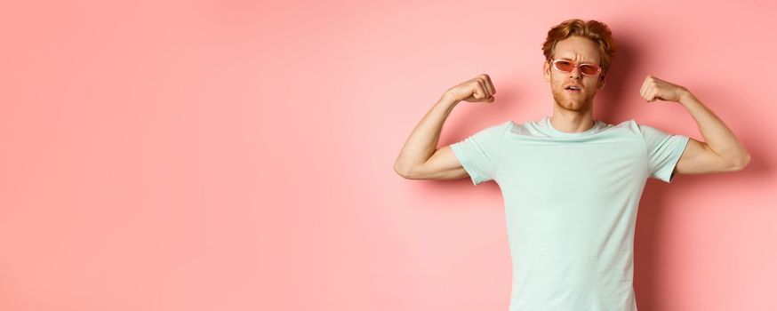 Confident young man with red hair, wearing summer sunglasses and t-shirt, showing strong and fit body muscles, flex biceps and staring cool at camera, pink background. Workout concept