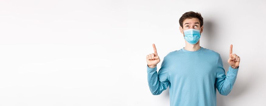 Coronavirus, health and quarantine concept. Curious guy reading banner on top, looking and pointing fingers up, standing in medical mask over white background.