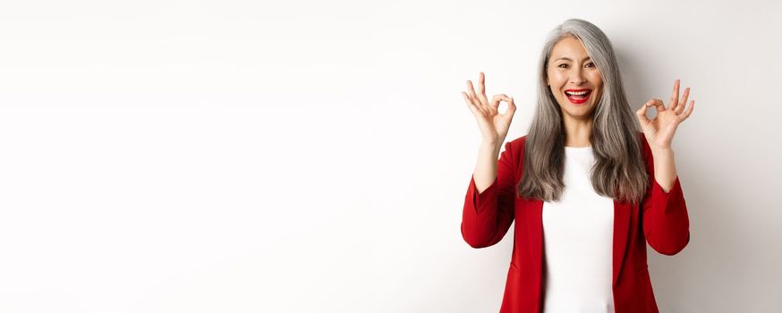 Beautiful senior asian woman in red blazer, smiling and showing okay signs, approve and say yes, standing over white background.