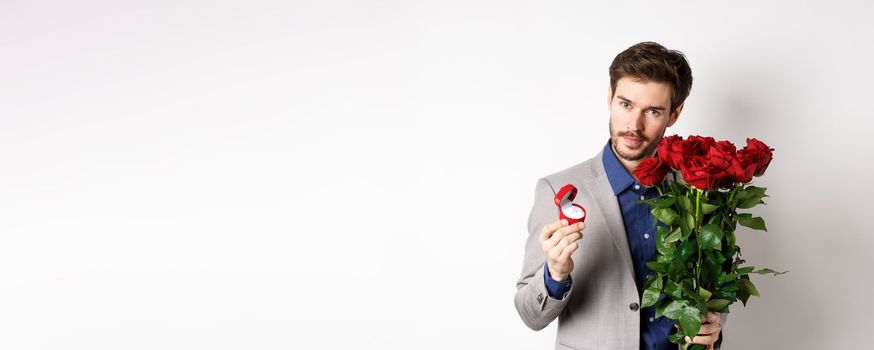 Handsome man in suit, showing engagement ring and looking romantic at camera, standing with red roses over white background. Valentines day and love concept.