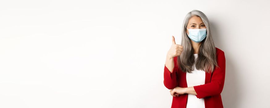 Coronavirus and business concept. Asian female entrepreneur in face mask looking cheerful, showing thumb-up in approval, praising using protective measures at work.