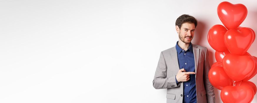 Lovers day. Charming young man with beard, wearing fancy suit, pointing finger at heart balloon surprise for Valentines day, standing over white background.