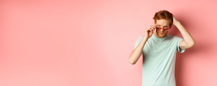 Handsome young redhead man in sunglasses, brushing hair with hand and looking smug and confident at camera, standing over pink background.