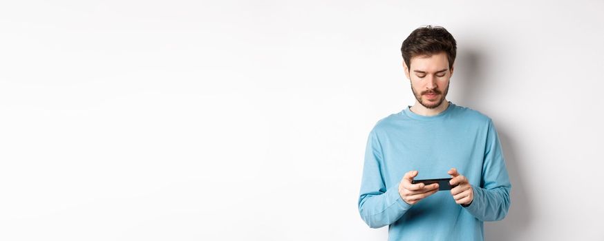 Male model playing video game on smartphone, looking serious at mobile screen, standing in sweatshirt over white background.