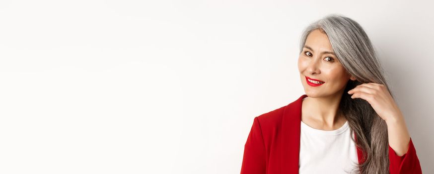 Beauty and haircare concept. Close up of elegant asian senior woman showing shiny and healthy grey hair, smiling and looking aside, white background.