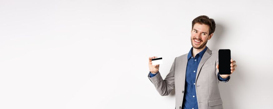 Online shopping. Successful businessman holding plastic credit card and showing mobile screen, smiling satisfied, making money, white background.