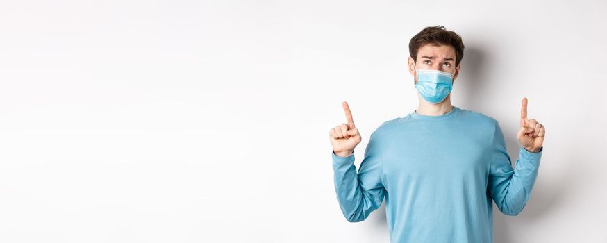 Coronavirus, health and quarantine concept. Skeptical and doubtful man in medical mask pointing, looking up, feeling indecisive, standing over white background.