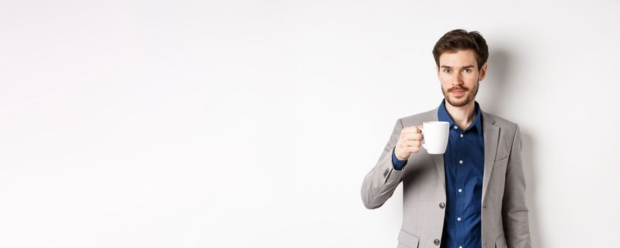 Handsome businessman in suit drinking coffee or tea from office mug, standing against white background.