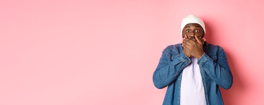 Shocked african-american man cover mouth and staring at camera, standing speechless against pink background.