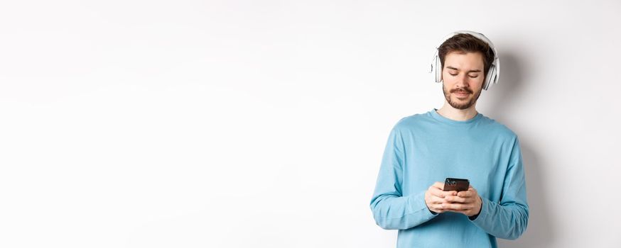 Modern young man in blue sweatshirt put on music on smartphone, listening songs in wireless headphones, white background.