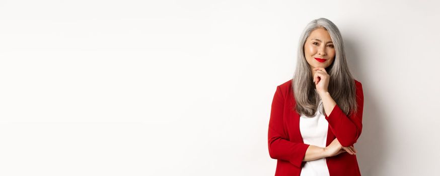 Business concept. Asian mature businesswoman smiling pleased, looking thoughtful, having an idea, standing in red blazer over white background.