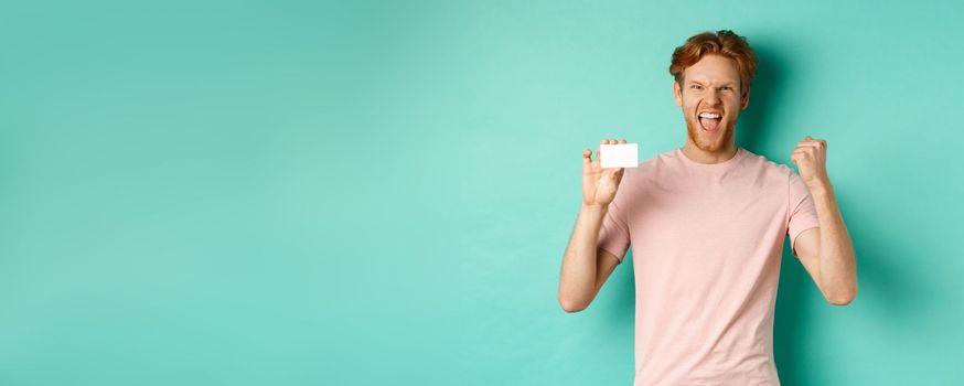 Cheerful young man triumphing, making fist pump to celebrate success, showing plastic credit card, winning prize from bank, turquoise background.