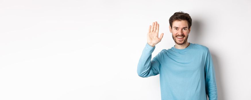 Cheerful young man with beard saying hello, looking friendly and waving hand to greet you, standing over white background.