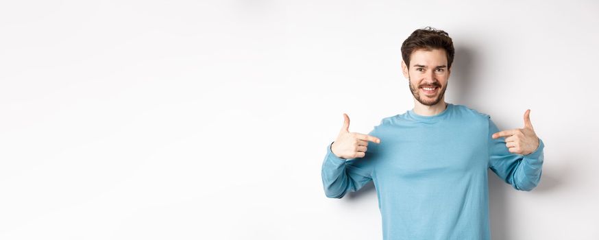 Handsome bearded man pointing fingers at center, self-promoting and looking confident, standing over white background, smiling at camera.
