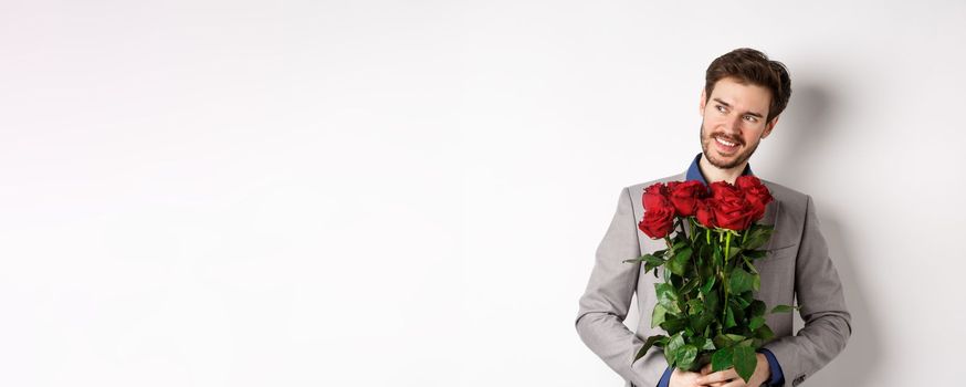 Charming smiling man going on romantic date, wearing suit, holding bouquet of roses and looking left, standing on valentines day with gift on white background.