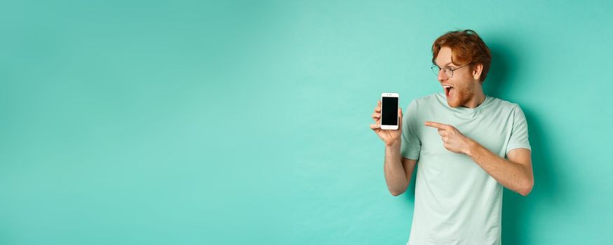 Check this out. Handsome redhead guy in glasses pointing finger at blank smartphone screen, showing online promotion, standing amazed over turquoise background.