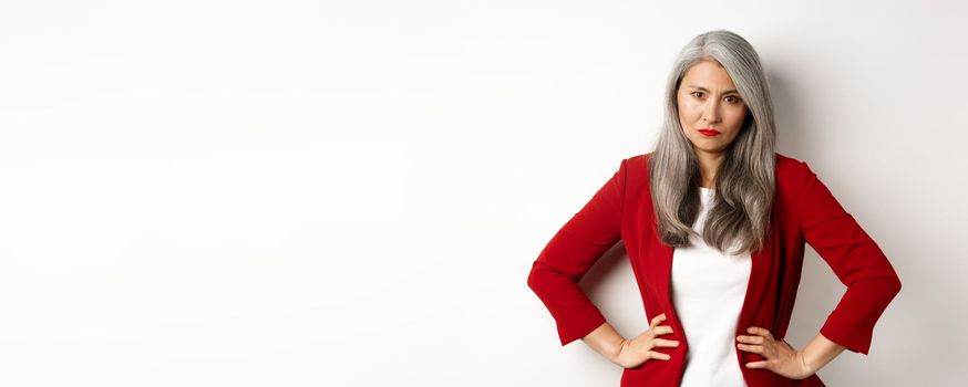 Angry asian senior businesswoman scolding employee, holding hands on waist and staring disappointed at camera, white background.