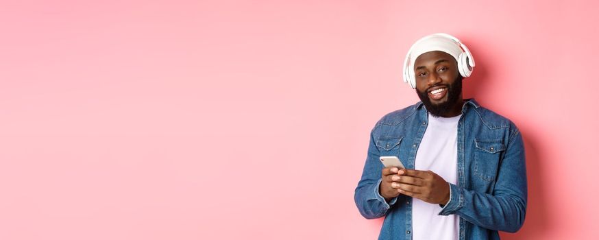 Handsome hipster guy in headphones smiling satisfied at camera, listening music in headphones, using mobile app, standing over pink background.