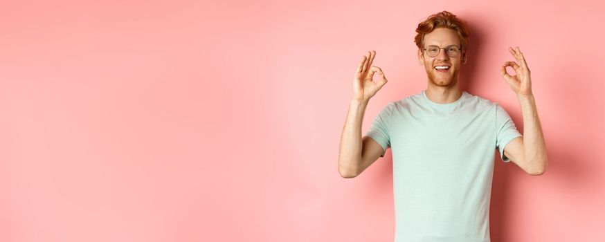 Satisfied caucasian man with red hair, wearing glasses and t-shirt, showing okay gestures and smiling, saying yes and approving something, standing over pink background.