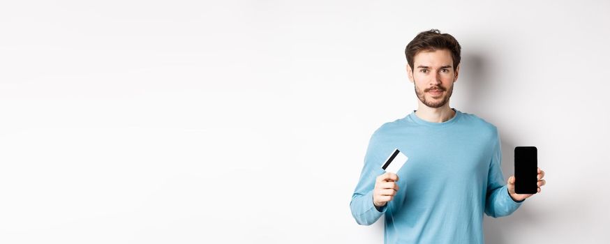 Handsome caucasian man showing empty smartphone screen and plastic credit card, standing over white background.