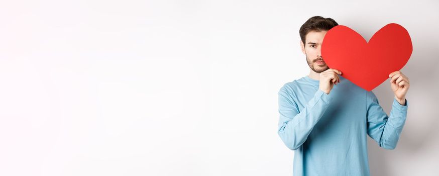 Young caucasian man holding Valentines day romantic heart, cover half of face with cutout and smiling, searching for love date, white background.