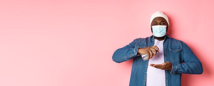 Covid-19, lifestyle and lockdown concept. Smiling african-american man in face mask cleaning hands with sanitizer, using antiseptic and looking at camera, pink background.