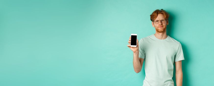 Skeptical male model with red hair and glasses, showing mobile screen and frowning disappointed, dislike application, standing over turquoise background.