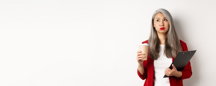 Tired asian female office worker holding clipboard and paper cup, drinking coffee and exhaling with exhausted face, standing over white background.