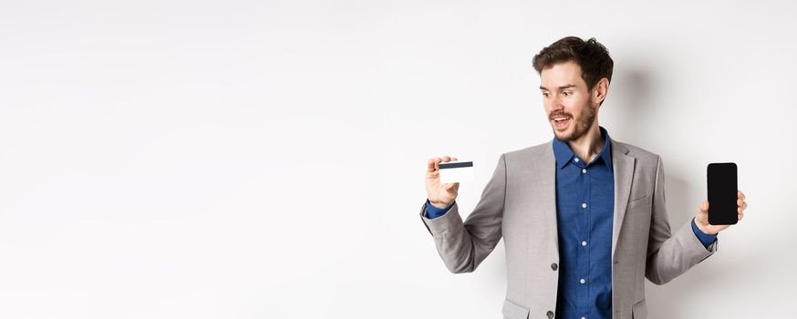 Online shopping. Smiling business man in suit showing plastic credit card with empty smartphone screen, standing against white background.