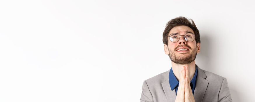 Nervous hopeful man in glasses and suit begging god, asking please and shaking hands in pray, white background.