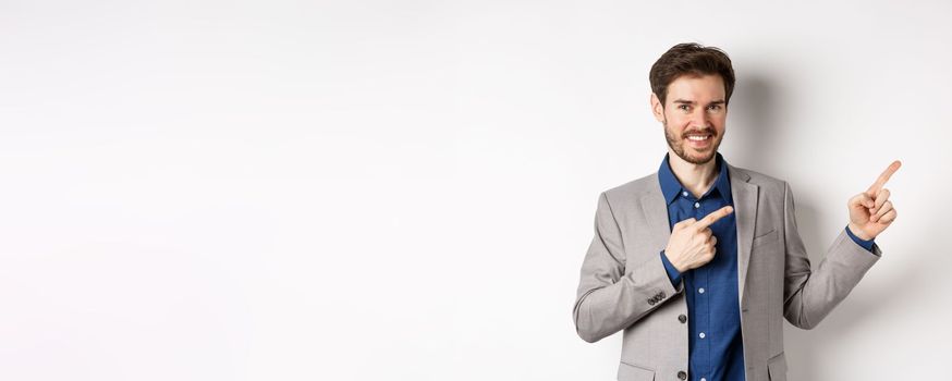 Handsome smiling man in business suit pointing fingers at upper right corner, showing logo, advertising company, standing on white background.